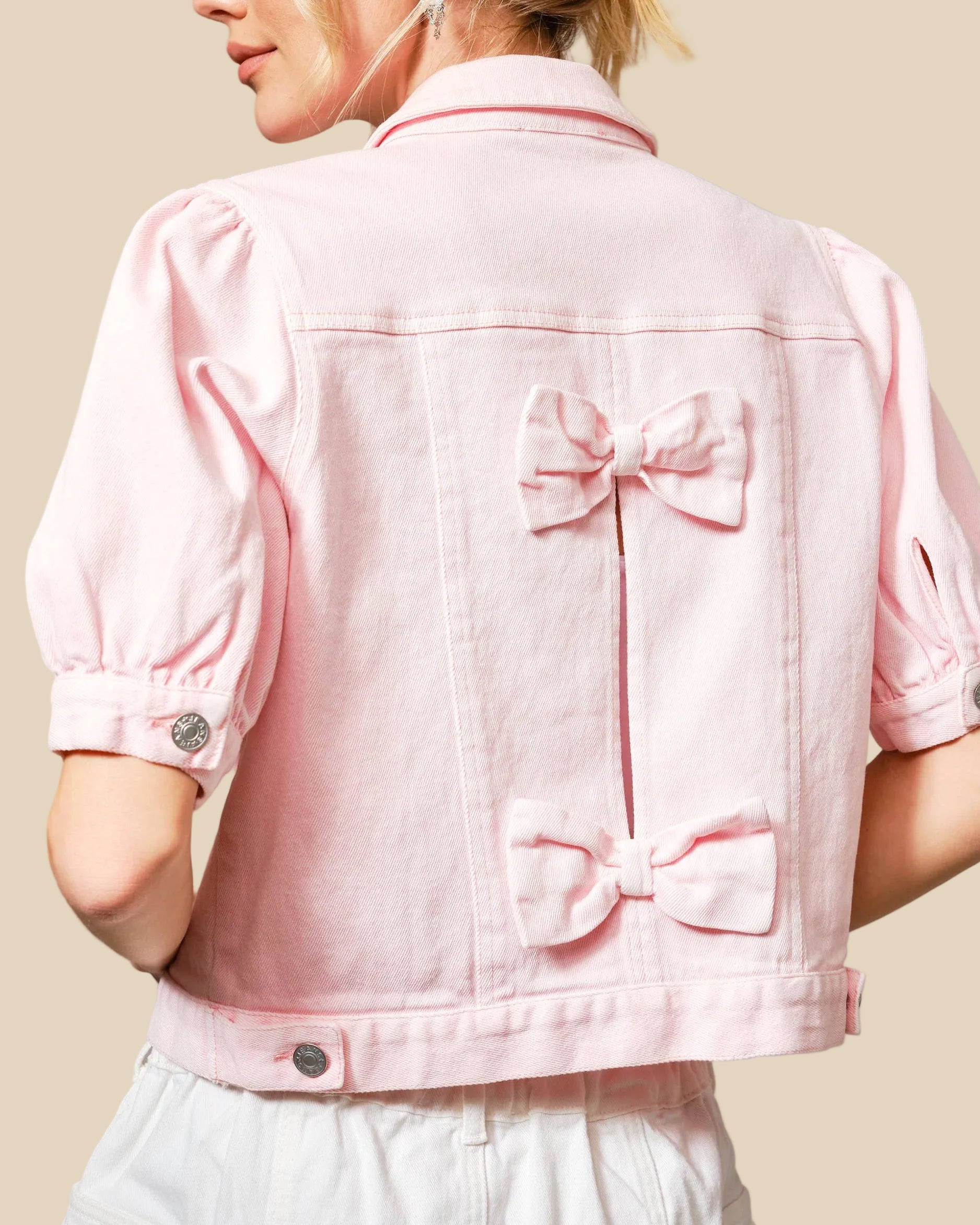 Pink denim jacket with bow details worn by a person on a gray background