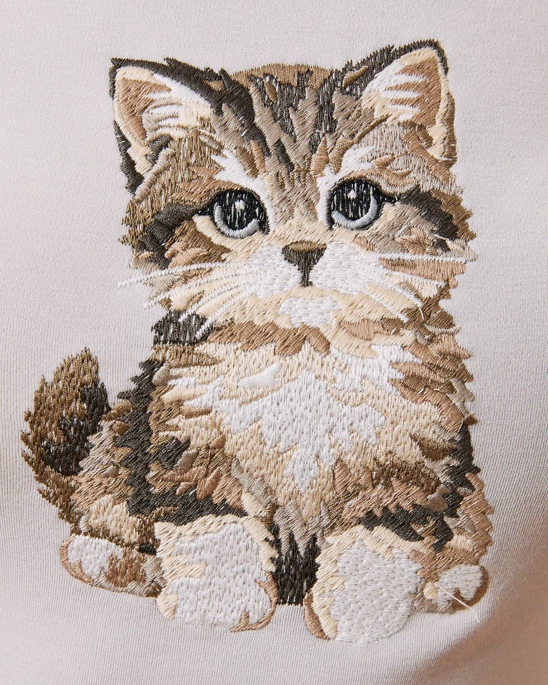 A close-up of a beige or cream-colored fabric featuring a detailed, hand-embroidered kitten design in shades of brown, beige, and white.